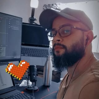 Pessoa de barba, óculos e boné cinza, sentada em frente a uma mesa de trabalho com dois monitores, um laptop, teclado mecânico e um microfone de estúdio. A iluminação é suave, com uma luminária ao fundo. Um coração pixelado em tons de laranja e vermelho aparece na imagem, dando um toque descontraído.